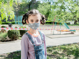 A little girl in a protective mask stands near a playground in the street during the pandemic of coronavirus and Covid - 19