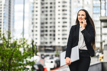 A young woman in smart casual wear with a laptop is walking on the cases, she is talking on the smartphone