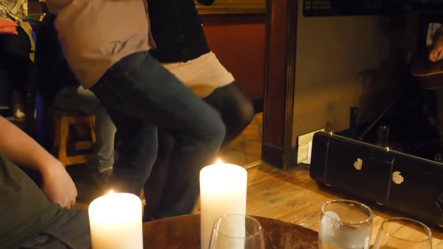 Excited couple dancing traditional dance energetically stamping floor in the Irish candle lit pub while other people watching and guitar playing in the background; handheld camera shot.