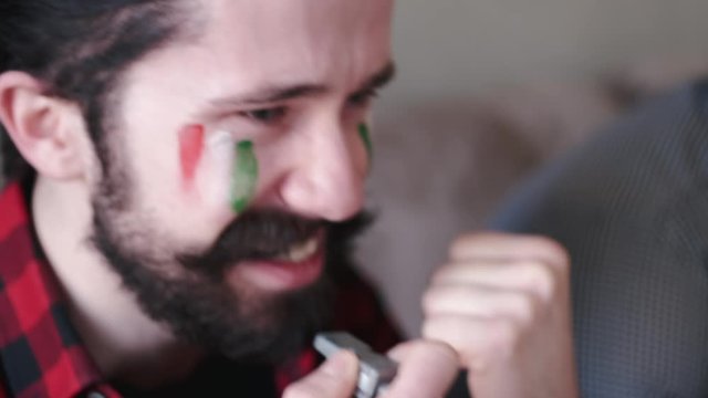 Man painted Italian flag on face . He has dark hair and beard. Person is watching tv at home.