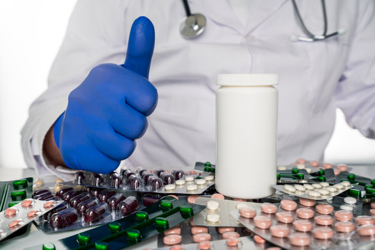 Doctor in white coat sits infront of pill boards scattered on table giving thumbs up next to blank prescription bottle. Hospital, vaccination and medicine concept.