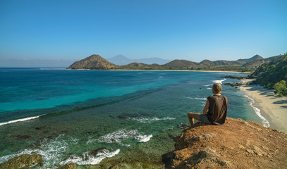 Blond man sitting and enjoying the views to undiscovered beach, hills and Rinjani Volcano seen in Sumbawa island