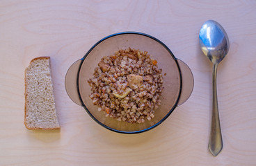 Buckwheat porridge with meat, seasoned with pieces of carrots and onions in glass brown transparent bowl, standing on wooden surface, next to spoon and piece of bread. Concept: simple village food.