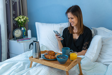 Happy young woman using mobile phone while having tasty breakfast on a tray in bed