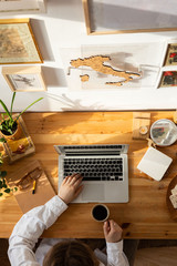 Wooden desktop top view, woman designer hands using laptop and holding a cup of coffee. Freelancer working place - houseplants, cookies, picture on the wall, sketchbook, vintage book. Work from home.