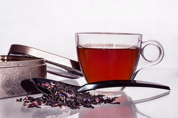 A Cup of tea on a glass table. Next to the Cup is a black spoon with tea leaves and a metal jar.