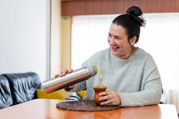 Fototapeta na wymiar Middle aged woman drinks the traditional Argentine yerba mate while laughing with an expression of happiness in a living room of a house. He wears a gray sweater.
