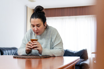 Middle aged white woman drinks traditional Argentine yerba mate on a wooden table with a living...
