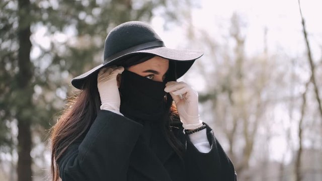 A beautiful young woman in black style is out for a walk. As it is quarantine period so she is wearing a protective mask and gloves in a park. She is strolling through paths.
