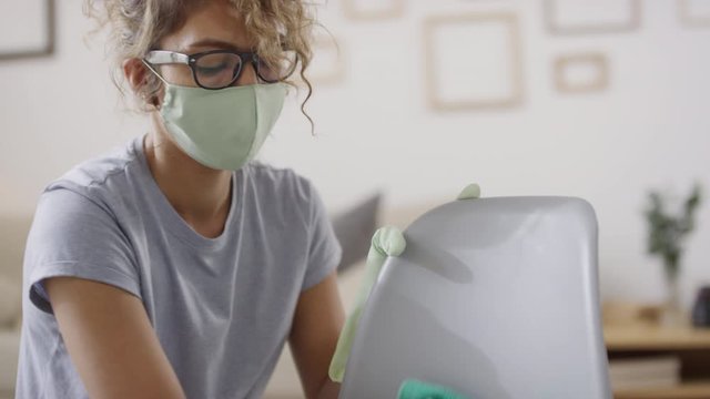 Tilt down shot of young woman in face mask and gloves cleaning chair surface at home