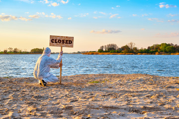 man in protective suit and gloves sets warning sign with inscription closed along the sand beach of the river. Contaminated water, quarantine, virus outbreak, crime scene concept
