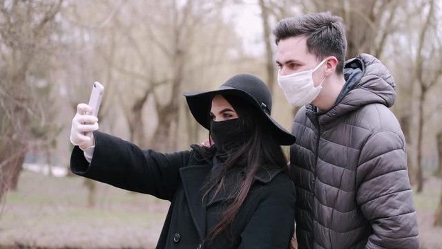 The young couple are out for a date in a park. It is quarantine time so they are wearing protective equipment. They are taking photos on a smartphone.