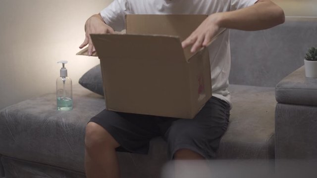 Asian male unboxing cardboard box, on sofa at home, mail receiver unpack small carton parcel package, covid-19 crisis, package delivery online purchase delivery, unpacking package sit on sofa at home