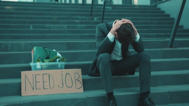 Worried young man sitting on stairs head off with box of personal stuff and poster with the inscription Need Job got fired due to coronavirus crisis unemployed depression economy slow motion