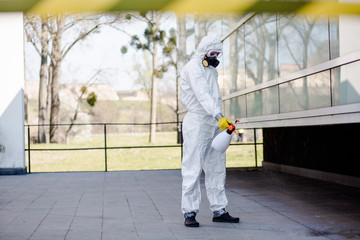 Man wearing an NBC personal protective equipment suit, gloves, mask, and face shield, cleaning the streets with a backpack of pressurized spray disinfectant water to remove covid-19 coronavirus.