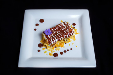 Tasty sweet dessert. A piece of chocolate biscuit cake with white air cream, drops of milk chocolate with thin slices of hazelnut. Black background