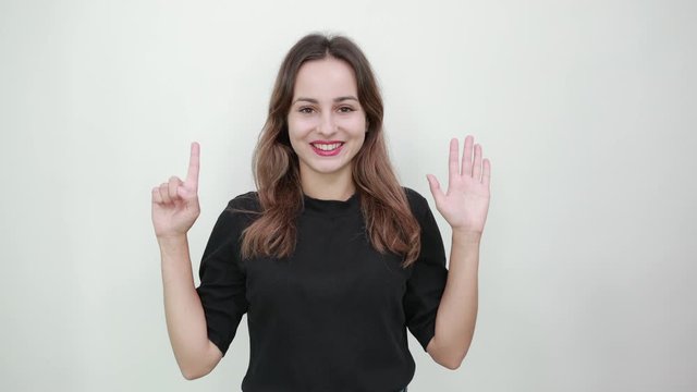 Cute Young Brunette Woman In Black T-Shirt, Blue Jeans With Belt On Gray Background, Happy Girl Smiles And Shows Six Fingers