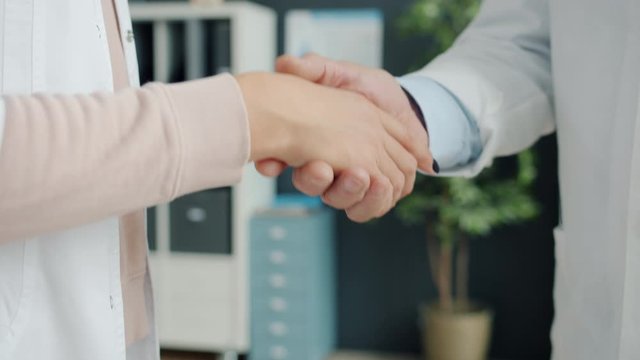 Close-up of male and female shaking hands wearing medical gowns indoors cooperating in hospital. Doctors cooperation and moden medicine concept.