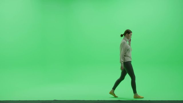 Green Screen Chroma Key Studio: Beautiful Young Woman Wearing Stylish Casual Clothes Uses Smartphone, Holding and Talking on the Mobile Phone Walks Across Room left to Right. Side View Camera Shot