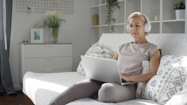 Tracking shot of attractive middle aged woman sitting on sofa at home, listening to music with headphones and typing on laptop