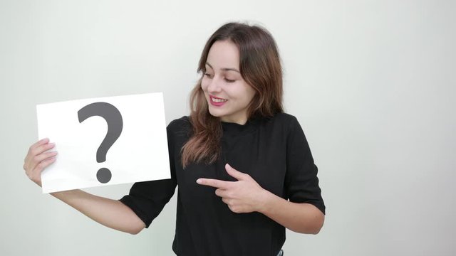 Cute Young Brunette Woman In Black T-Shirt, Blue Jeans With Belt On Gray Background, Happy Girl Smiling Holding Piece Of Paper With Question Mark In Hand And Pointing Index Finger In The Direction