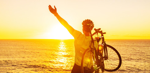 Success, achievement, accomplishment and winning concept with cyclist mountain biking. Happy MTB woman cycling raising arms lifting bike by sea during sunset cheering and celebrating at summit top.