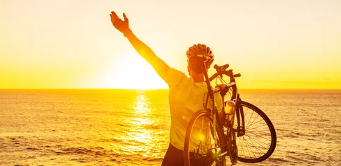 Foto op Plexiglas Success, achievement, accomplishment and winning concept with cyclist mountain biking. Happy MTB woman cycling raising arms lifting bike by sea during sunset cheering and celebrating at summit top. © Maridav