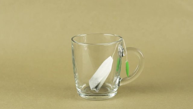 black tea bag with green label is placed in glass transparent mug and filled with boiling water isolated on beige background slow motion