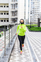 A girl with a protective mask on the face with headphones on her head is running in the city street. Morning jog during quarantine