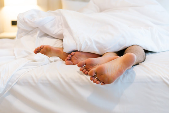 Close up of male and female feet on a white bed. The concept of loving couple having sex under white sheets in the bedroom. Sensual and intimate moment of lovers. Focus on male foot.