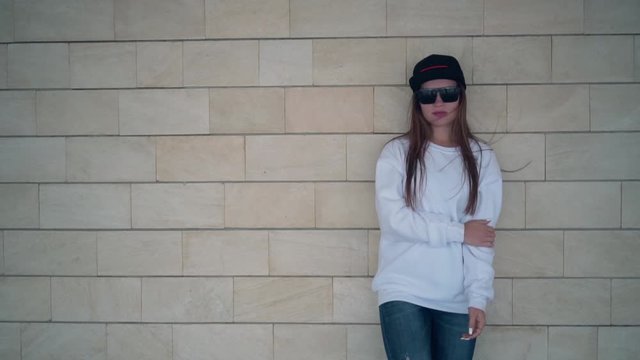 A young and sexy girl with long hair in sunglasses and a white sweatshirt and jeans stands against a beige wall of bricks, blocks. Stylish young girl stands on a background of a brick wall.