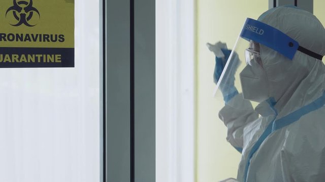 worker wearing personal protective equipment or ppe including mask, suit and face shield using disinfectant to clean window of isolation unit to prevent covid-19 or coronavirus spreading in hospital
