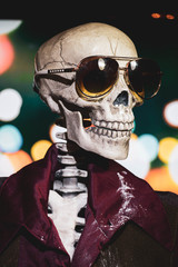 A skeleton dressed in a 1970's era collared shirt and vest, with sunglasses. White cocaine powder on his face and collar. Set against a scene of blurred city lights.