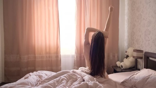 Young topless girl with long dark hair wakes up in a large bed in the bedroom
