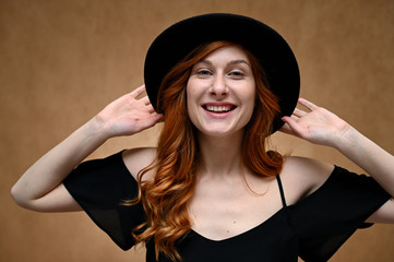 Portrait of a young happy woman with long red hair in a black hat on a beige background. Model with a smile posing.