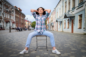 Brunette funny pretty girl sitting on chair in Central part of old city. Walk in downtown. Portrait of girl on street