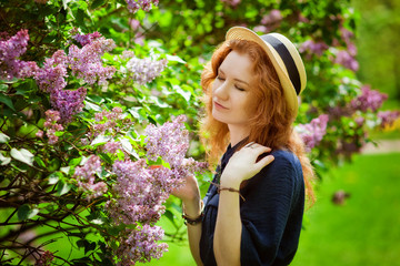 A beautiful red-haired girl in a straw hat in a Lilac garden. The girl breathes in the scent of blooming lilac. Gardening. Spring time.
