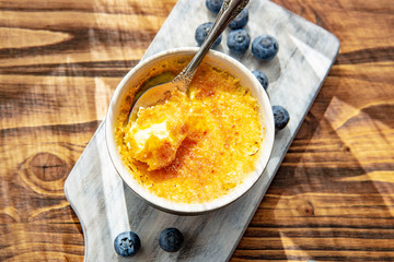 Creme brulee or catalana - traditional French vanilla cream dessert with caramelised sugar, blueberries on rustic white wooden board, table with sunny light. Sweet dessert -custard. Copy space.