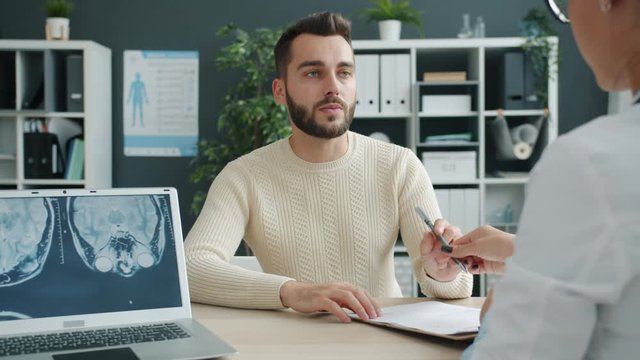 Worried patient young man is looking at mri results on laptop screen then signing medical document talking to doctor driendly woman in hospital office.