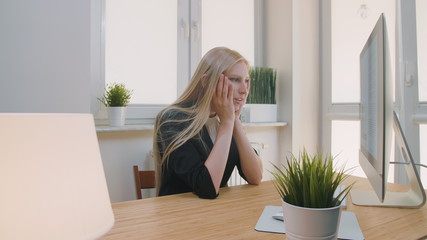 Blond sad female in elegant clothes clutching head after failure or bad news sitting in light office at desk with computer