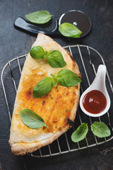 Freshly baked calzone pizza with green basil leaves on a metal cooling rack, vertical shot on a dark brown stone surface