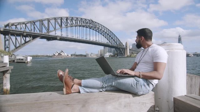 Man Using Laptop on Bench by Sydney Harbour Bridge. Wide Dolly Left