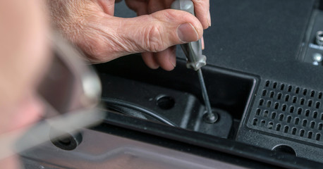 old man in glasses holds wrinkled hand on grey metal screwdriver repairing TV set at home