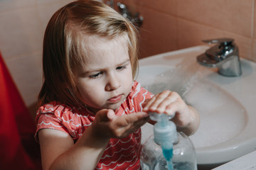 Cute little toddler girl press bottle dispenser with liquid soap for washing her hands in a bathroom. Hygiene concept showing prevention of infection and viruses such as flu and covid-19