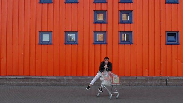 Happy customer man rides an empty shopping cart along the wall of mall store center outside and gives a thumbs up. Discounts and sales concept. Funny and amusement moment
