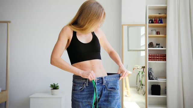 Beautiful young woman measures waist with meter to track progress after workout and fitness fat loss program. Fit model tries on oversize old jeans. Reach goal to look like magazine cover