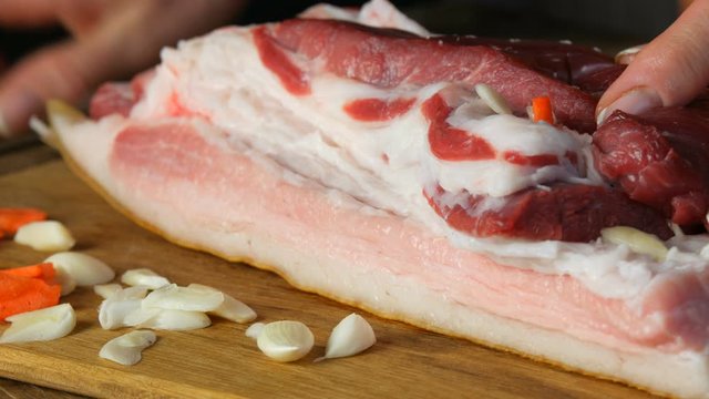 Female hands stabs with a knife and stuffed seasonings with slices of garlic and carrots a piece of fresh raw meat pork steak or bacon in a rustic style on wooden kitchen board