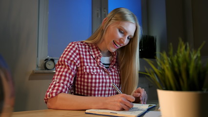 Lively young blond woman in casual checkered shirt, sitting at wooden desk with notebook holding metal pen and looking at camera smiling at night in dark room.