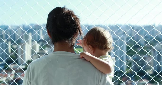 Mother holding baby overlooking city at apartment balcony with safety net window