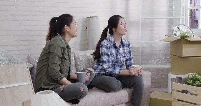 slow motion sweet asian lesbian discussing place furniture for building new family house. two young girls sitting on sofa point out window with urban view look from apartment relax laugh chatting